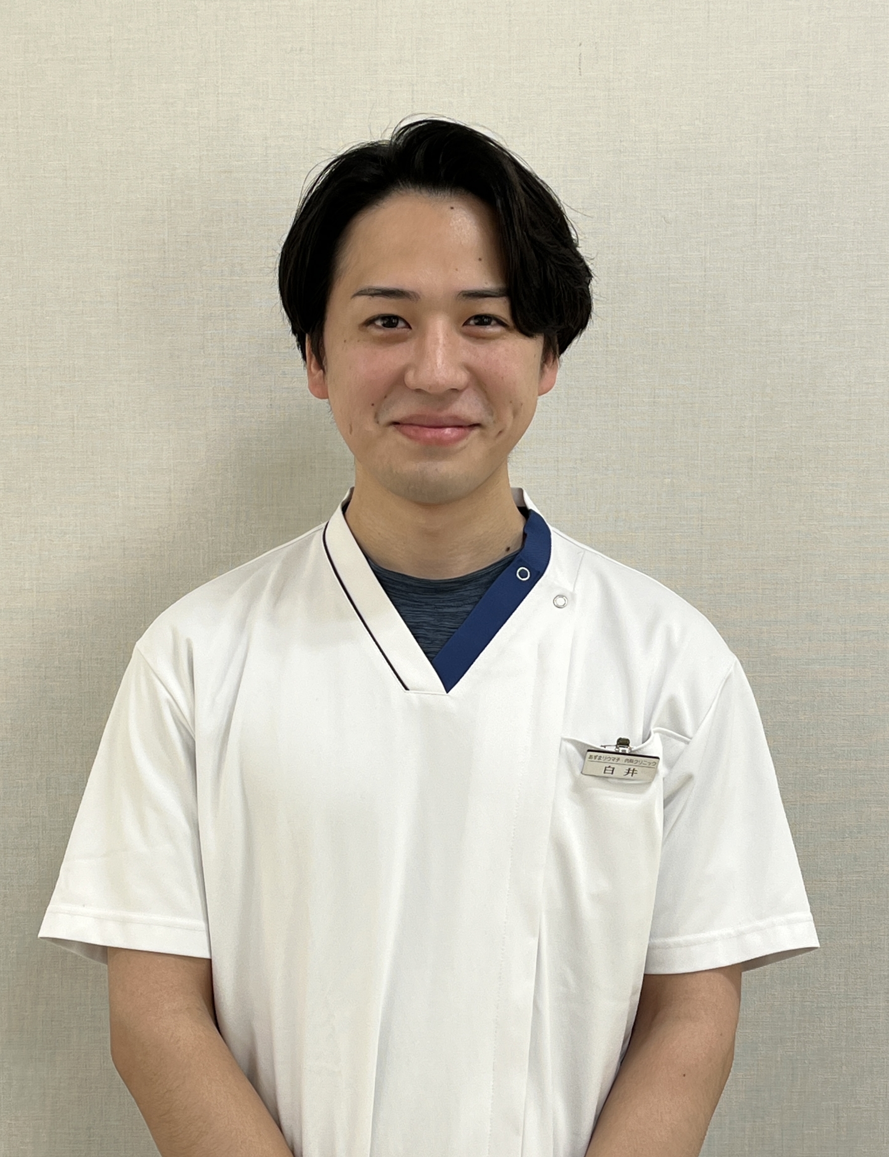 Smiling healthcare professional in a white scrubs-style uniform with a name badge standing against a beige wall.