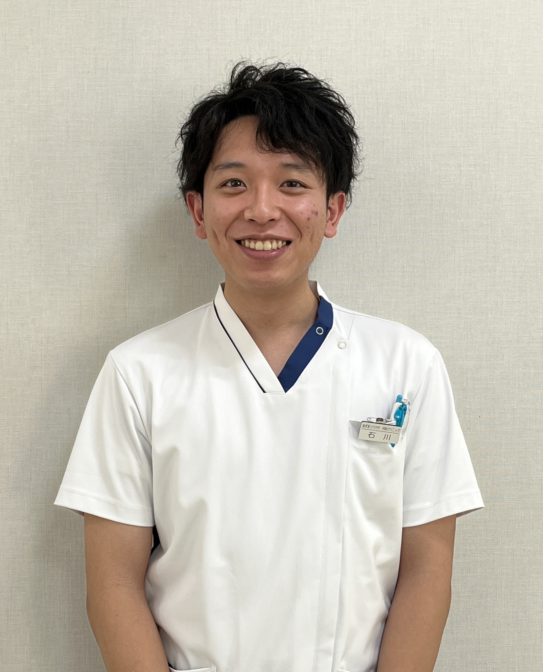 Young man in white medical scrubs smiling at the camera against a beige wall.