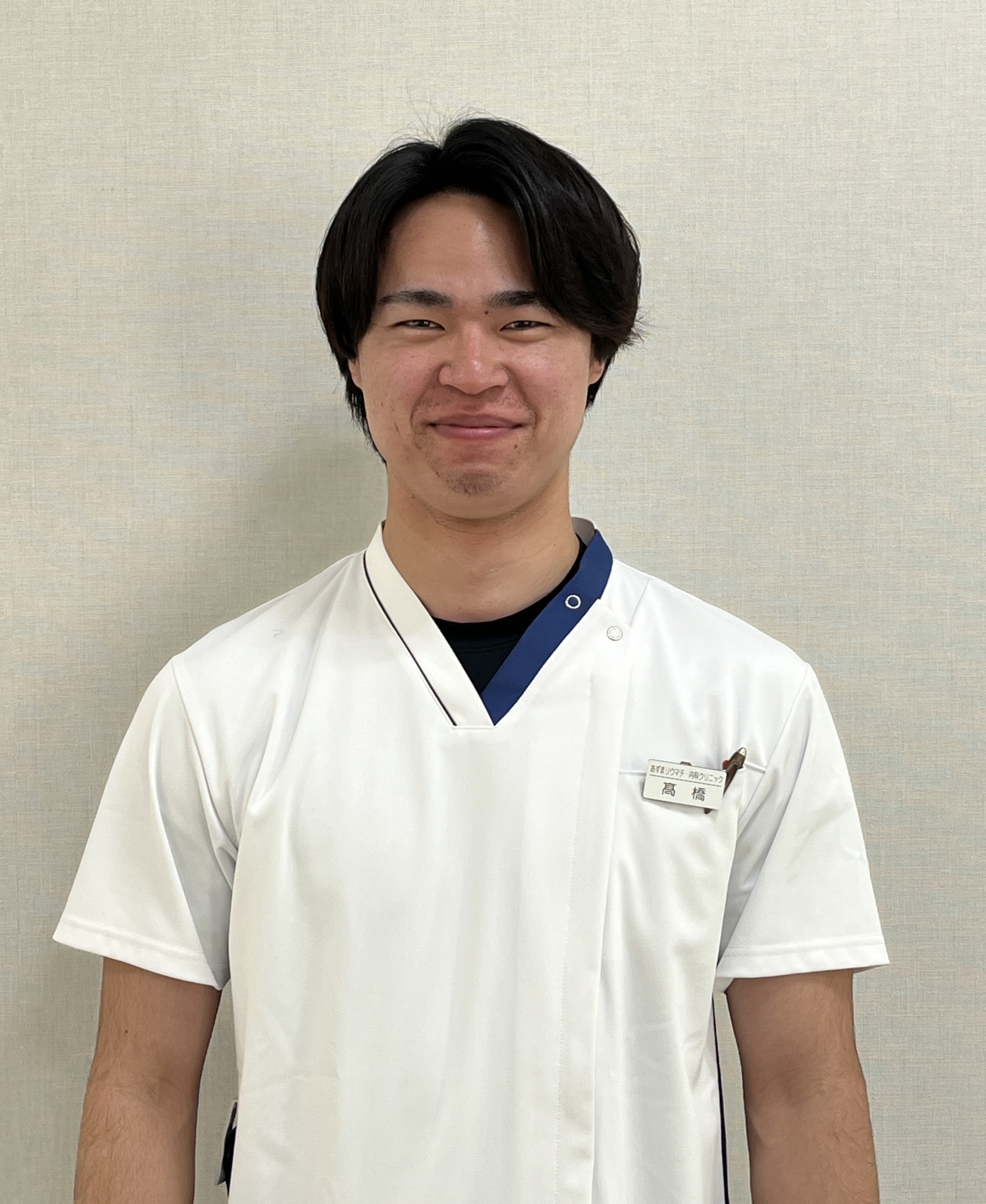 Young man in a white medical uniform with a name badge, standing against a beige wall.